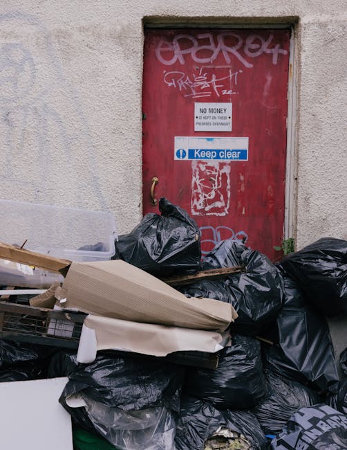 A collection of black trash bags and discarded packaging materials, including cardboard boxes and plastic wrapping, are piled outside a building against a textured, beige wall. The accumulation is near a red metal door that has graffiti and multiple signs attached, including a white 'No Money' notice and a blue 'Keep Clear' sign. The bags appear to contain waste or unwanted household items, indicative of a recent clearance or removal process. The scene suggests an area awaiting collection, with objects possibly ready for disposal or preparation for home relocation services. The setting is outdoors on a pavement or loading area, with natural daylight illuminating the scene, and some small green plants are visible near the base of the wall, adding to the contextual environment. This image represents home clearance or waste removal, processes often associated with professional removals such as those by Man With a Van Blackheath.