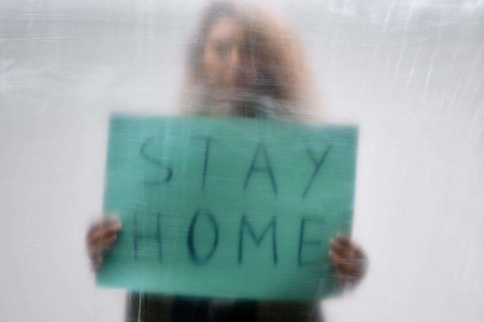 A person standing behind a frosted or foggy glass surface, holding a rectangular sign that reads 'STAY HOME' in large, clear letters. The individual is slightly blurred, with only their hands and part of the sign clearly visible, suggesting a focus on the message rather than their identity. The background appears to be neutral and out of focus. This image emphasizes the importance of home safety and is related to community health messages, which can be associated with home relocation or moving services such as those provided by Man With a Van Blackheath, especially during times of increased awareness about health precautions in moving or packing contexts.