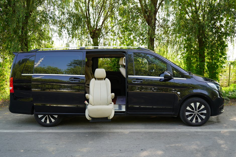 A black multi-passenger van parked on a residential street side with tall green trees and foliage in the background. The side sliding door is open, revealing a beige leather interior with a single front seat pulled out and positioned at the van's entrance, as if for loading or unloading. The van's interior visible includes a folded third-row seat, various packed cardboard boxes wrapped in plastic and bubble wrap, and furniture coverings. The scene shows the ongoing process of home relocation, with furniture and packing materials prepared for transport. The van, equipped with a sliding side door for easy access, is parked on an asphalt surface beneath a bright sky, supported by the professional moving company Man With a Van Blackheath, specialising in removals and furniture transport services, adhering to proper loading procedures for efficient packing and moving.