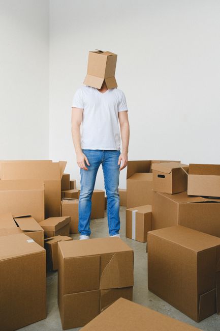 A person standing indoors surrounded by numerous unpacked cardboard boxes of varying sizes, some sealed with packing tape and others open with flaps folded back. The individual is wearing a white t-shirt and blue jeans, with a cardboard box placed on their head, concealing their face. The scene appears to be part of a home relocation process, with the boxes arranged on the floor and against the plain, light-colored wall in a well-lit room. This image reflects the packing and moving stage of house removals, with the environment suggesting an ongoing move managed by a professional moving service such as Man With a Van Blackheath. Equipment such as trolleys, straps, or vans are not visible in this image.