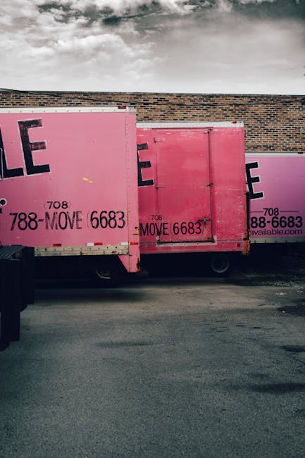 Image showing three large pink commercial moving containers positioned along a paved area beside a brick wall under a cloudy sky. The containers, used for home relocation and furniture transport, are made of metal with visible locking mechanisms and some signage including a phone number and the word 'MOVE'. The containers are placed to facilitate loading or unloading activities, with one container partially overlapped by another. The environment appears to be an outdoor space adjacent to a building, ready for loading furniture and belongings for a house removal service, as offered by Man With a Van Blackheath. The overall scene captures the preparation phase of packing and moving operations, emphasizing the logistics involved in professional relocation services.
