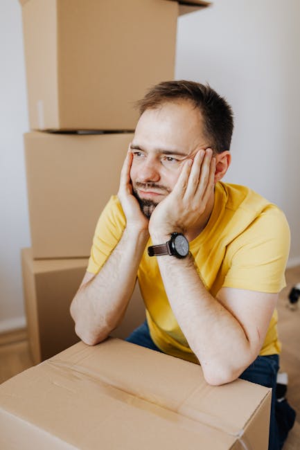 A person standing indoors surrounded by numerous unpacked cardboard boxes of varying sizes, some sealed with packing tape and others open with flaps folded back. The individual is wearing a white t-shirt and blue jeans, with a cardboard box placed on their head, concealing their face. The scene appears to be part of a home relocation process, with the boxes arranged on the floor and against the plain, light-colored wall in a well-lit room. This image reflects the packing and moving stage of house removals, with the environment suggesting an ongoing move managed by a professional moving service such as Man With a Van Blackheath. Equipment such as trolleys, straps, or vans are not visible in this image.