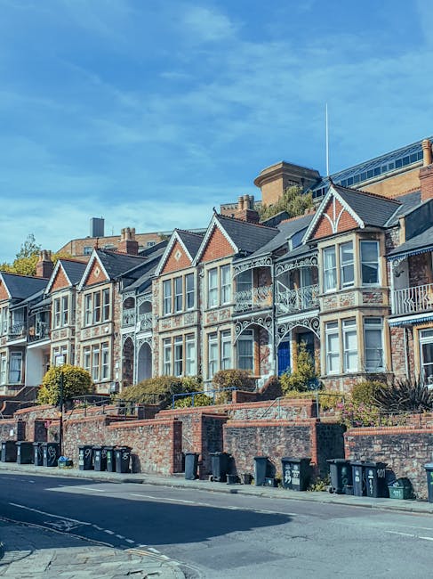 A row of Victorian-style terraced houses with brick façades, decorative white ironwork, and bay windows, situated on a residential street in Blackheath. The houses are built on an elevated walkway with brick retaining walls and small gardens with shrubs and plants. The street below has black wheelie bins lined up along the pavement. The bright blue sky with some scattered clouds overhead indicates clear weather. This image captures a typical urban residential scene relevant to house removals and home relocation activities, as handled by Man With a Van Blackheath, including the process of loading or arranging belongings for transport in a moving service context.