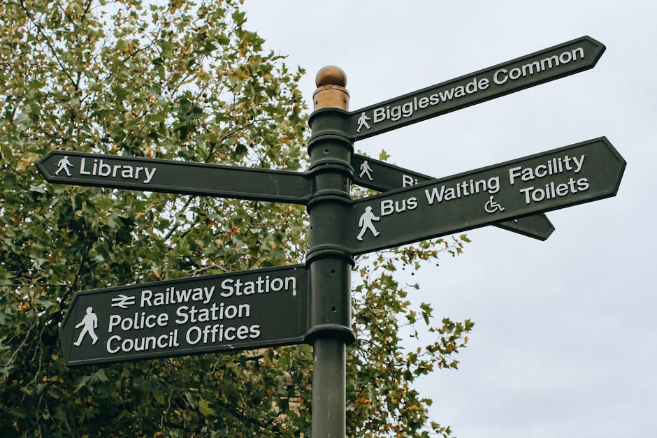 A black metal directional signpost situated outdoors against a backdrop of leafy green trees and a cloudy sky. The signpost features six rectangular panels with white text and icons, pointing in different directions: one towards Biggleswade Common, another towards a library, a third towards a bus waiting facility and toilets with a wheelchair accessible symbol, a fourth towards railway station and police station with a train icon, and a fifth towards council offices. The panels are mounted on a central pole topped with a rounded finial. The image captures the signage as part of an urban environment, possibly near residential or commercial areas, highlighting navigation for pedestrians and visitors. Man With a Van Blackheath is associated with removals and relocation services, indicating that the signpost might be relevant for guidance during home removals, furniture transport, or packing and moving activities in the Blackheath area.