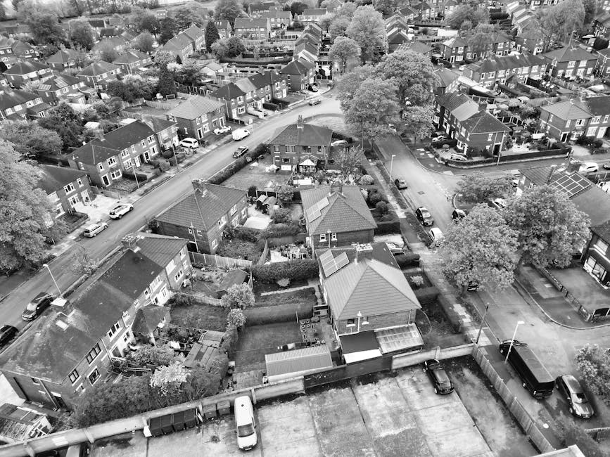 An aerial black-and-white photograph of a residential neighborhood in Blackheath shows multiple houses, some with pitched roofs and gardens, arranged along curved and straight roads. Several cars are parked along the streets and in driveways. In the foreground, the rooftops of terraced and semi-detached houses are visible, with back gardens showing garden sheds, small trees, and hedges. Large trees are scattered throughout the area, providing shaded areas in front and rear gardens. A narrow alleyway separates some properties, with fenced boundaries defining individual gardens. The scene captures the quiet, well-established suburban environment suitable for home relocation and furniture transport, with clear pathways for moving logistics. Man With a Van Blackheath specializes in removals, supporting efficient house moves like those depicted, where careful loading and transport of furniture and boxes are integral to the process, often involving van loading areas seen at the street level in the image.