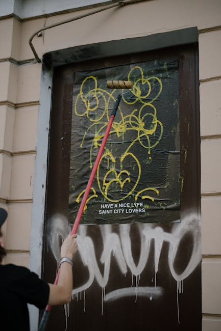 Image showing a person using a long-handled roller brush to paint or clean a black, plastic-covered window or door on the exterior of a building in an urban environment. The building has beige or cream-colored siding, and a portion of a person's arm and shoulder are visible on the left side of the image, suggesting active work. The window or door is decorated with yellow graffiti-style artwork featuring abstract, flower-like shapes, and there is a printed message reading 'HAVE A NICE LYFE SAINT CITY LOVERS' beneath the artwork. Nearby, there is a white graffiti tag partially visible at the bottom of the door or window. The setting appears to be during daylight with natural lighting. This scene is related to home or business premises, possibly indicating exterior maintenance or creative street art, with the visible context aligning with property remodelling or urban art projects, which can relate to furniture or materials involved in moving or renovation activities.