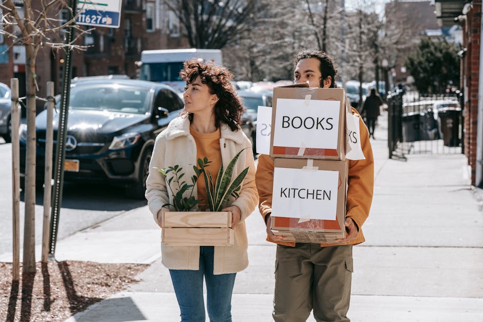 Two individuals are walking along a residential street, each carrying boxes and items related to a house move. The woman on the left has curly brown hair and is wearing a beige fleece coat over an orange top, holding a small wooden crate filled with potted plants. The man on the right, with dark curly hair, is dressed in a tan jacket and khaki pants, carrying two stacked cardboard boxes labeled 'BOOKS' and 'KITCHEN.' The boxes are secured with clear tape and have printed labels indicating their contents, suggestive of packing and moving preparations. They are walking on a paved sidewalk adjacent to buildings with brick facades, with parked cars visible in the background, including a black van or hatchback near a tree. The scene is illuminated by daylight, with some snow on the ground, indicating a cold season. This image reflects the process of home relocation, involving furniture transport and packing, consistent with services offered by Man With a Van Blackheath during a house removal.