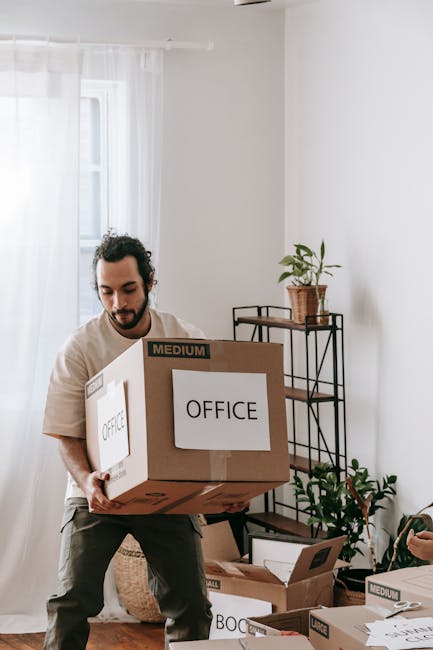 An individual with dark, curly hair and a beard, wearing a red long-sleeved shirt, stands indoors against a plain, light-colored wall, smiling gently while holding a cardboard box labeled 'OFFICE' in 