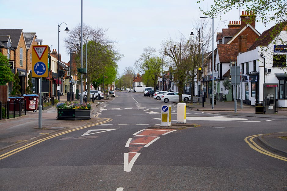 A street view in Blackheath showing a designated loading and unloading zone with parking suspensions, marked with road signs and road markings. The area features a small traffic island with a blue directional arrow and yellow bollards preventing vehicle entry. Surrounding buildings include residential and commercial properties, with vehicles parked along the street. Trees line the sidewalks, and streetlights are visible at intervals. The scene is well-lit with natural daylight, illustrating an urban environment suitable for home relocation and furniture transport logistics. Man With a Van Blackheath frequently operates within such areas, assisting with removals, packing, and transporting household furniture through designated loading bays and parking suspensions for efficient moving services.