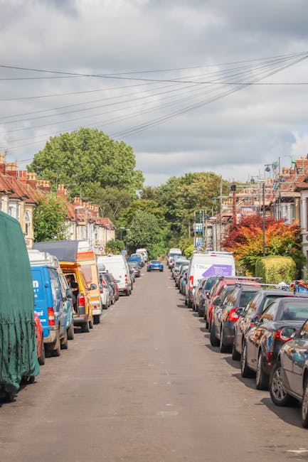 A street view in Blackheath showing a designated loading and unloading zone with parking suspensions, marked with road signs and road markings. The area features a small traffic island with a blue directional arrow and yellow bollards preventing vehicle entry. Surrounding buildings include residential and commercial properties, with vehicles parked along the street. Trees line the sidewalks, and streetlights are visible at intervals. The scene is well-lit with natural daylight, illustrating an urban environment suitable for home relocation and furniture transport logistics. Man With a Van Blackheath frequently operates within such areas, assisting with removals, packing, and transporting household furniture through designated loading bays and parking suspensions for efficient moving services.