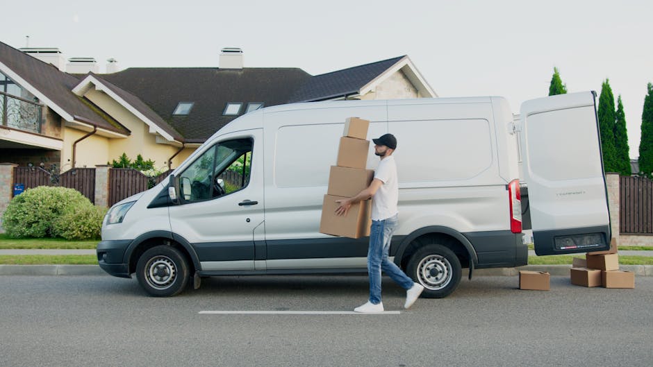 A man wearing a white t-shirt, jeans, and a black cap is seen outdoors in front of a white delivery van during daylight hours. He is carrying three cardboard boxes, stacking them as he walks alongside the van, which is parked on a residential street. Inside the van, visible through the open side door, are additional cardboard boxes and wrapped furniture pieces, all secured with packing materials such as plastic wrap and blankets. The van is positioned beside a paved pavement, with a row of modern houses featuring pitched roofs, dormer windows, and a garden with green shrubs and trees in the background. Several boxes are placed on the ground near the van's rear, indicating ongoing loading or unloading, part of the home relocation or furniture transport process managed by Man With a Van Blackheath, operating within the removals service category. The environment is well-lit, with natural daylight illuminating the scene, capturing the logistical steps involved in packing and moving household items.