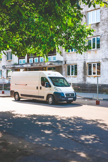 A white cargo van parked on a street in front of an apartment building during daylight hours, with the van positioned parallel to the pavement. The van features a sliding side door and a high roof, suitable for home relocation and furniture transport. Surrounding the vehicle are several delivery straps and blankets, indicating the loading process for a house move. The van is parked close to the building's entrance, which is partially visible behind a large leafy tree casting shadows on the street and van. The building appears to be multi-storey with visible windows decorated with snowflake and star patterns in blue and white, suggesting a festive or residential setting. The overall scene reflects professional removals activities, with a focus on efficient removal logistics, such as loading household items and furniture into the vehicle, consistent with services offered by Man With a Van Blackheath.