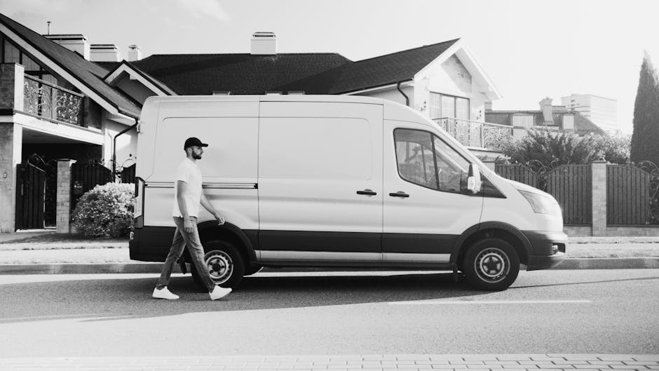 A man wearing a white t-shirt, jeans, and a black cap is seen outdoors in front of a white delivery van during daylight hours. He is carrying three cardboard boxes, stacking them as he walks alongside the van, which is parked on a residential street. Inside the van, visible through the open side door, are additional cardboard boxes and wrapped furniture pieces, all secured with packing materials such as plastic wrap and blankets. The van is positioned beside a paved pavement, with a row of modern houses featuring pitched roofs, dormer windows, and a garden with green shrubs and trees in the background. Several boxes are placed on the ground near the van's rear, indicating ongoing loading or unloading, part of the home relocation or furniture transport process managed by Man With a Van Blackheath, operating within the removals service category. The environment is well-lit, with natural daylight illuminating the scene, capturing the logistical steps involved in packing and moving household items.
