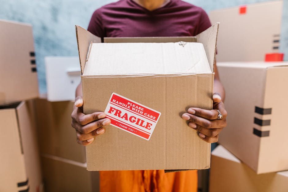 A person wearing a maroon top is holding a medium-sized cardboard box with both hands, positioned in front of their torso. The box is unsealed and appears ready for home relocation or packing and moving tasks. There is a red and white sticker on the front of the box that reads 'PLEASE HANDLE WITH CARE FRAGILE THANK YOU.' The background shows several other cardboard boxes of varying sizes, some sealed with black tape, stacked in a room likely used for packing and storing items prior to moving. The setting appears to be indoors, illuminated by natural or artificial light, with the nearby environment suggesting preparations for furniture transport and moving logistics, consistent with professional removals by Man With a Van Blackheath. The image emphasizes careful handling of delicate items during a house removal process, highlighting the importance of proper packing and transport procedures.