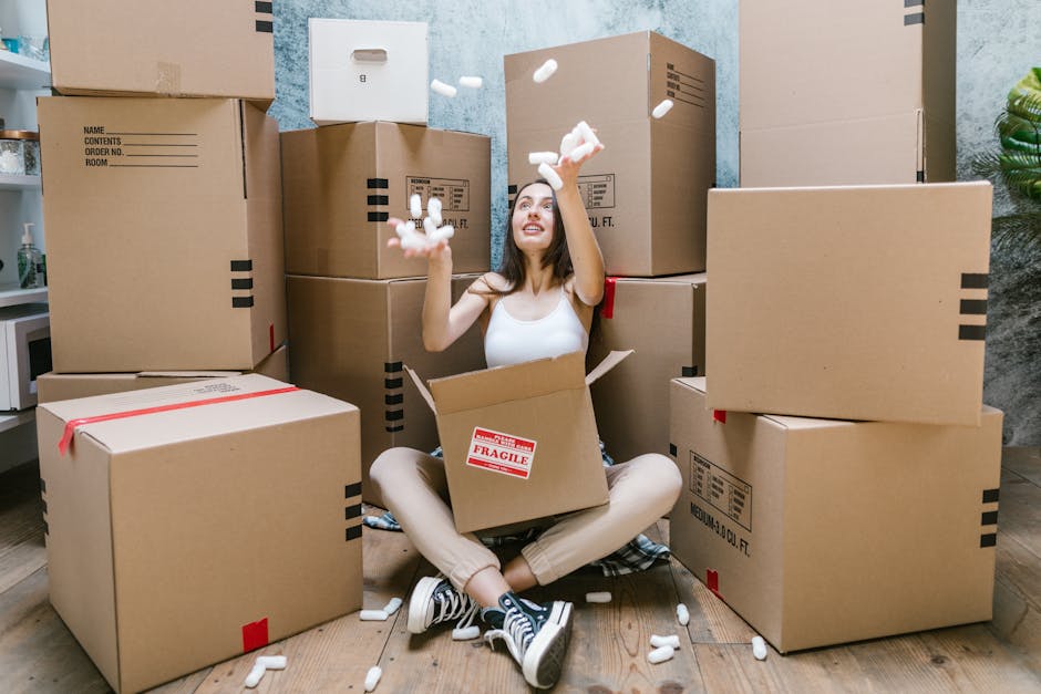 A woman sitting cross-legged on a wooden floor inside a property, surrounded by multiple large cardboard boxes of varying sizes, some sealed with red tape and others labeled with handling instructions, as she drops packing foam pieces from her hands. One box positioned in her lap has a red 'Fragile' sticker on it. The background includes textured light blue walls and a small part of a plant visible on the right side, indicating an interior space prepared for moving. This scene captures the packing and loading process involved in home relocation services provided by Man With a Van Blackheath, illustrating the careful handling and organized packing typical of furniture transport and moving logistics.