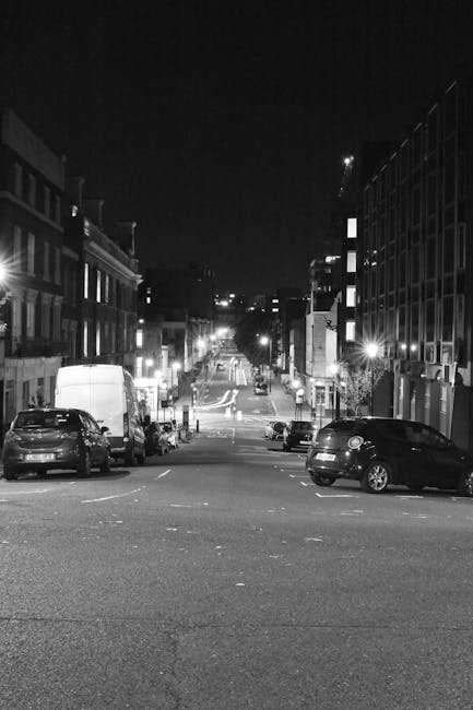 A black-and-white photograph taken at night showing a narrow urban street with parked cars along both sides, including a white van and a dark-colored hatchback in the foreground. The street is illumin