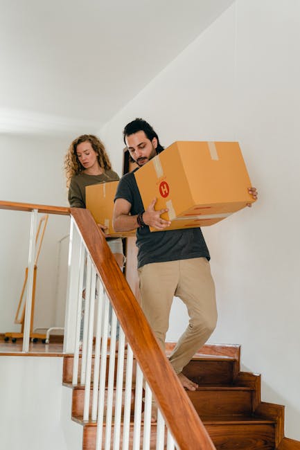Two movers from Man With a Van Blackheath are transporting cardboard boxes down a wooden staircase inside a residential property. The male mover in the foreground is holding a medium-sized box wrapped with packing tape, positioned at the bottom of the stairs, preparing to carry it further down. The female mover, standing behind him on the staircase landing, is holding a similar box with both hands, ready to assist with the home relocation process. The staircase features wooden steps with a white banister and railing, leading from an upper floor to a ground level. The interior has neutral-colored walls and ceiling, with natural light illuminating the scene. This loading and packing activity highlights the careful handling of household items during a furniture transport or moving service by [COMPANY_NAME] as part of their professional removals process.