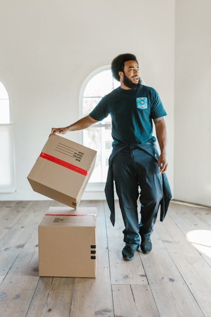 A man with a dark complexion, wearing a navy blue t-shirt with a printed design on the chest and dark trousers, is inside a bright, spacious room with light-colored walls and wooden flooring. He is holding a cardboard box with red tape in his right hand, preparing to place it on a second box positioned on the floor. The room features a large arched window allowing natural light to illuminate the space. The scene depicts the process of home relocation, with the man engaged in packing or unpacking activities as part of furniture transport and moving logistics, which aligns with services offered by Man With a Van Blackheath. The use of packing materials and careful handling of boxes suggest a professional moving or removals service focused on house moves, emphasizing the importance of organized loading and careful packing during a household move.
