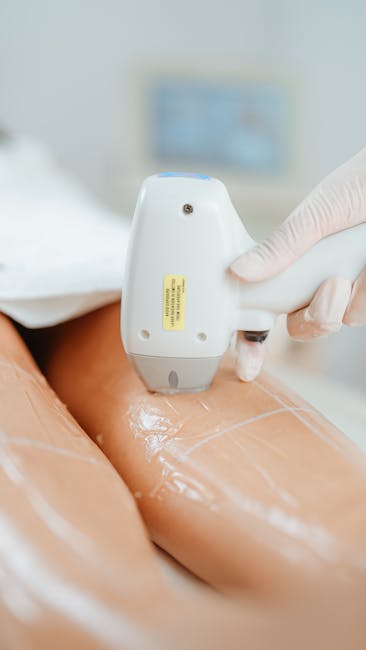 A close-up image showing a person wearing a white glove using a handheld ultrasound device on a person's thigh during a massage or treatment session in a clinical setting. The thigh is covered with a shiny, transparent gel to facilitate the procedure. In the background, there is a blurred monitor or screen, indicating a professional environment. The scene relates to treatment or therapy services, which may be part of a larger house removal or relocation service involving health or wellness checks prior to moving, with the equipment positioned on a clean surface within an indoor space. The image emphasizes the use of specialist tools, professional care, and preparation for a move, subtly aligning with the comprehensive process of packing, transport, and ensuring well-being during house removals, as described in the context of Blackheath removals.